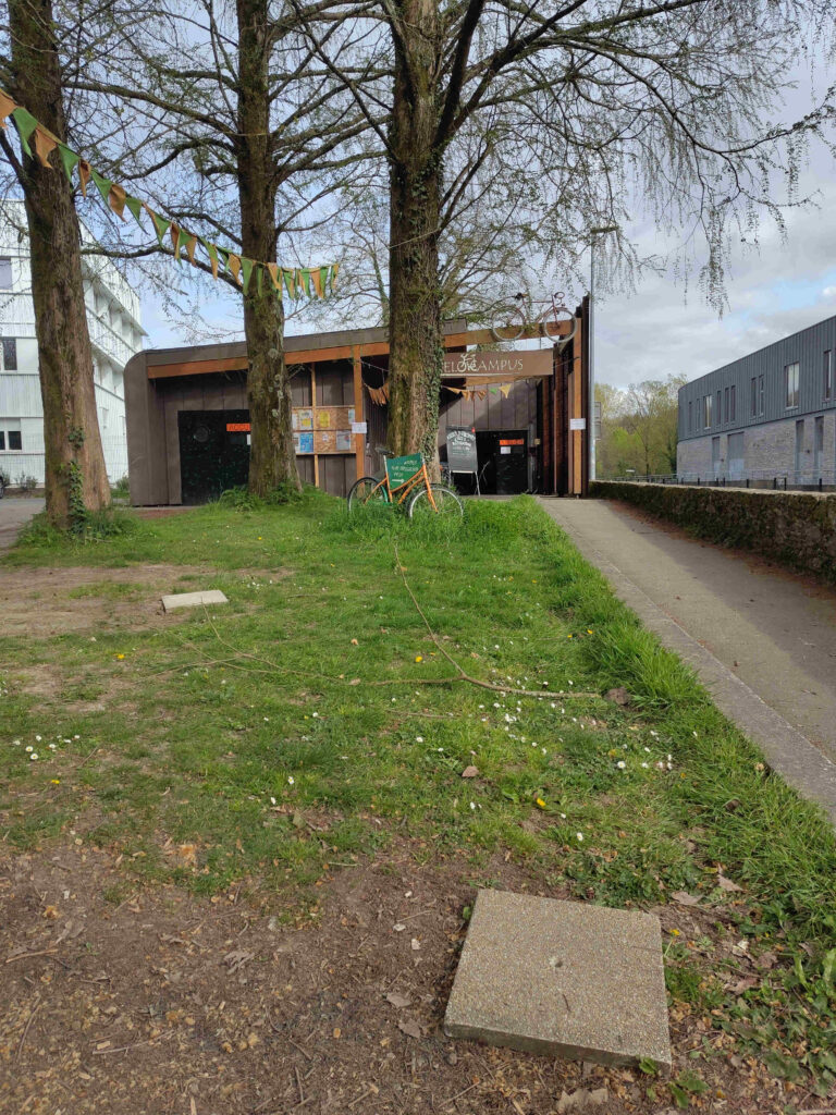 Photo devant l'atelier. Une rampe goudronnée passe sur le côté d'un parterre d'herbe, et mène à l'atelier ouvert